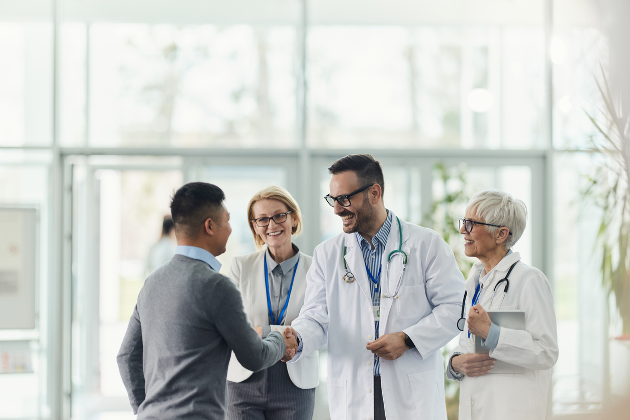 A healthcare professional greeting a visitor with a handshake while colleagues smile nearby.
