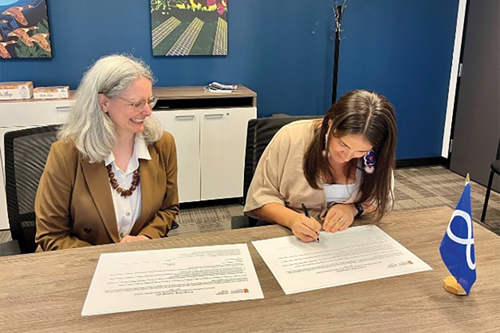 Two people sitting at a conference table, signing a partnership agreement document. One person is smiling while the other person is focused on writing. There is a small Métis Nation flag on the table in front of the documents.