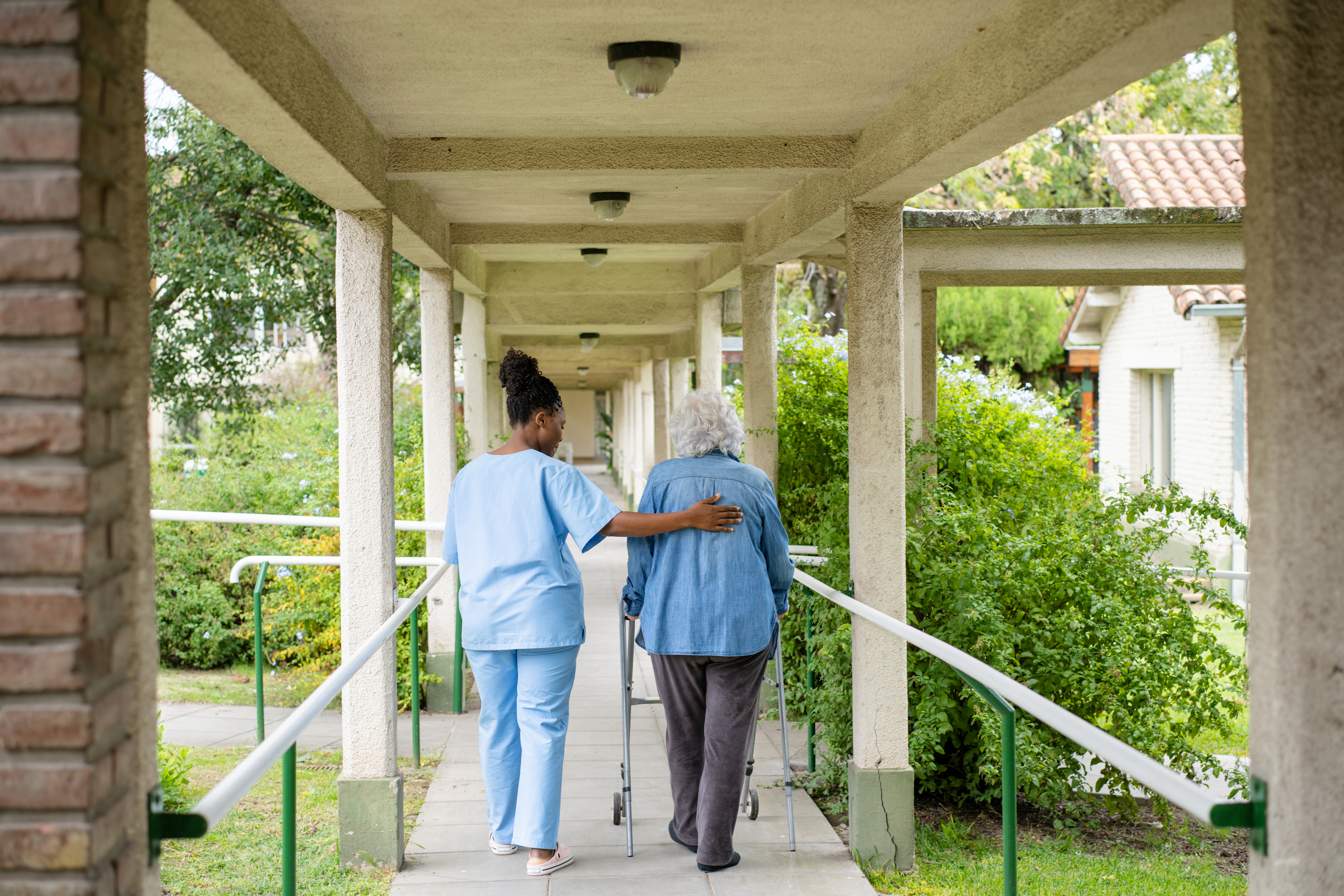 On a paved outdoor pathway, a caregiver gently supports a patient to walk along the path. The person is using a walking aide to support her. There are also guard rails on either side of the path.