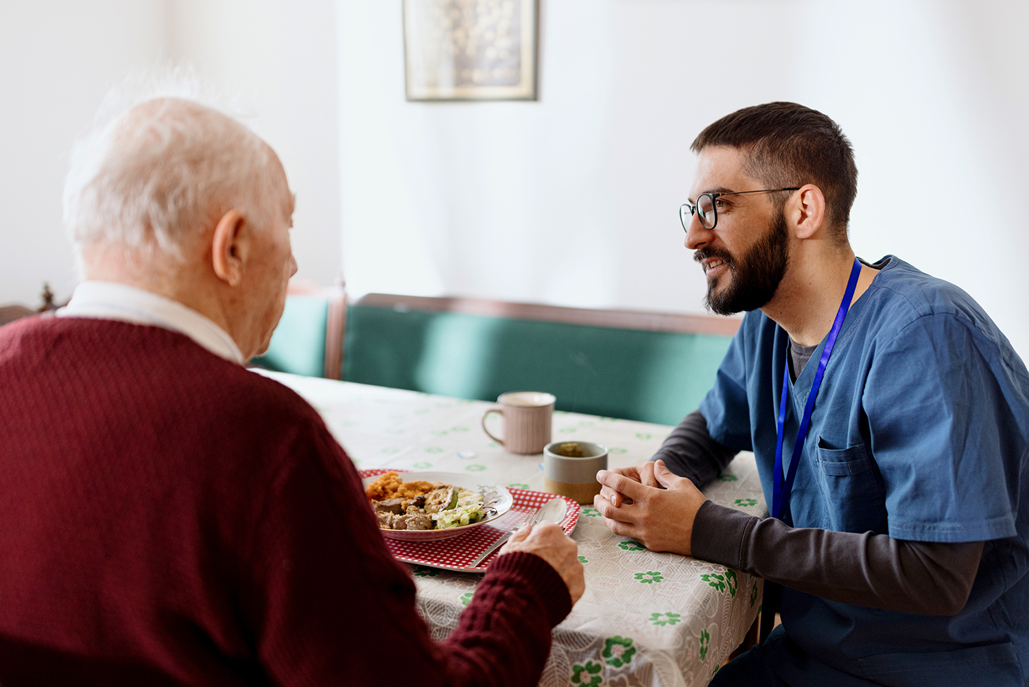 A male healthcare provider sits with his elderly male patient while he has breakfast and has a coffee.