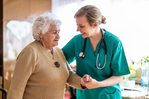 A female healthcare professional dressed in hospital attire assisting an elderly female patient to stand in a hospital room.