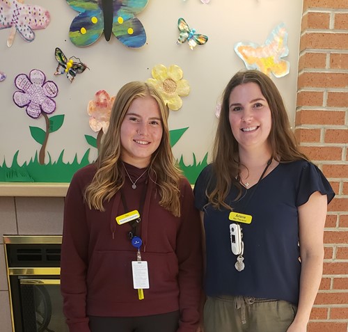 Two team members at The Pines home stand smiling in front of a wall decorated with colourful flowers and butterflies.