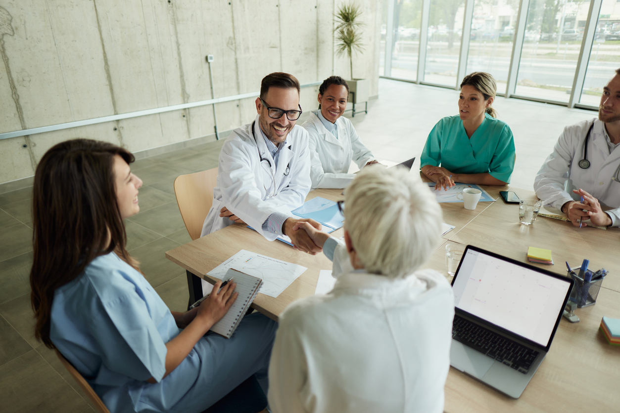 Healthcare professionals in a meeting, smiling as two colleagues shake hands across the table.