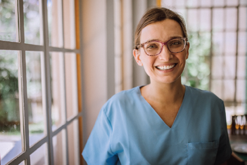 A female healthcare provider with glasses standing by a window smiling at the camera.